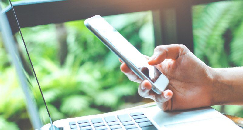 Hand holding a white smartphone, using it near a laptop, blurred green background.