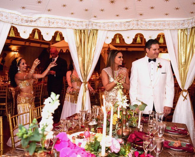 Couple at a wedding reception, guests applaud. Elaborate tent decor with gold accents and belly dancers.