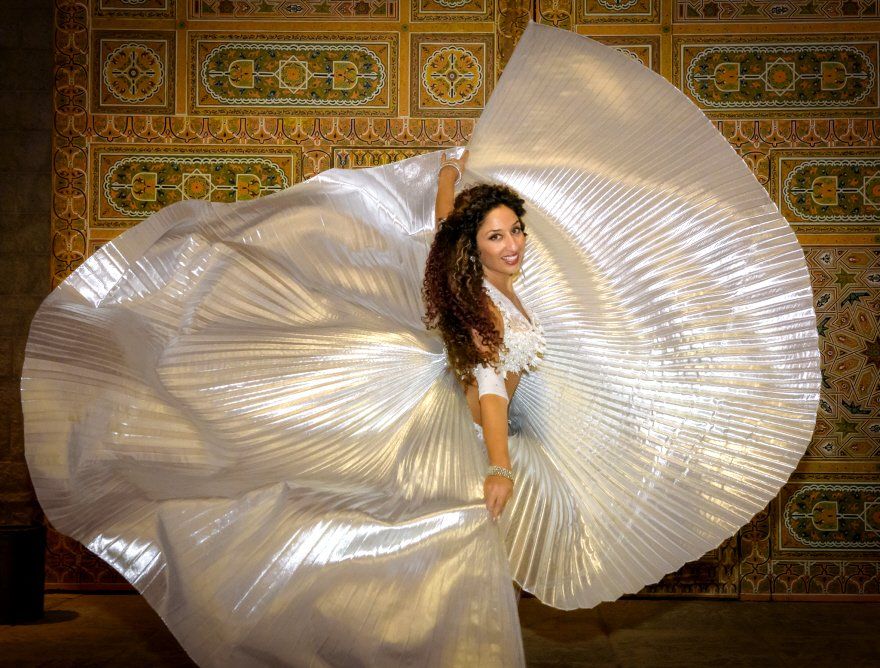 Belly dancer in white costume, arms outstretched, holding large, pleated fans, set against ornate, golden door.