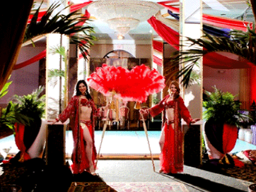 Two dancers in red outfits pose by a large red fan in a decorated room with palm trees.