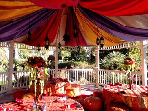 Gazebo decorated with colorful fabric drapes and lanterns, set with low seating and a table.
