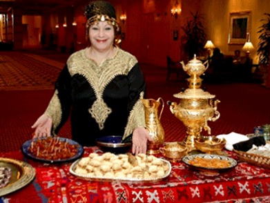 Woman in ornate outfit presents food at a table with golden samovar, tea set, and sweets. Red carpet backdrop.