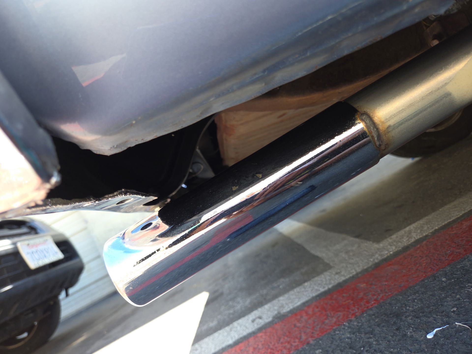 Close-up of a chrome car exhaust pipe under a bumper, with a tire visible in the background.