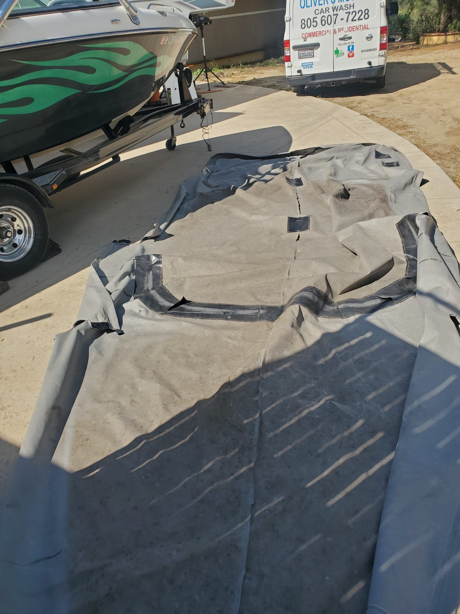 Deflated gray boat cover on a driveway beside a boat trailer and truck, with a storage bin in the background