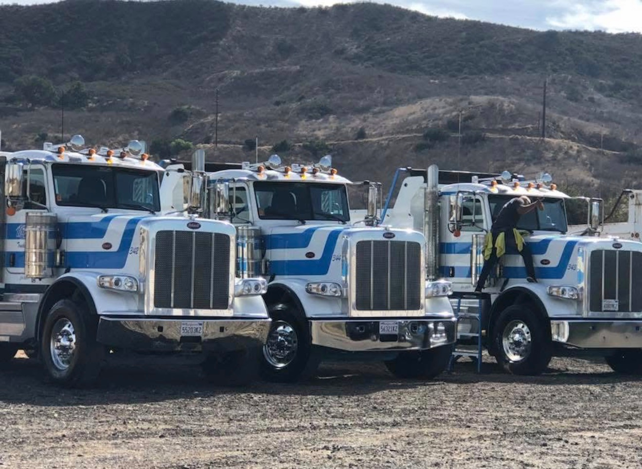 Three white-and-blue semi trucks parked in a row on gravel with hills in the background