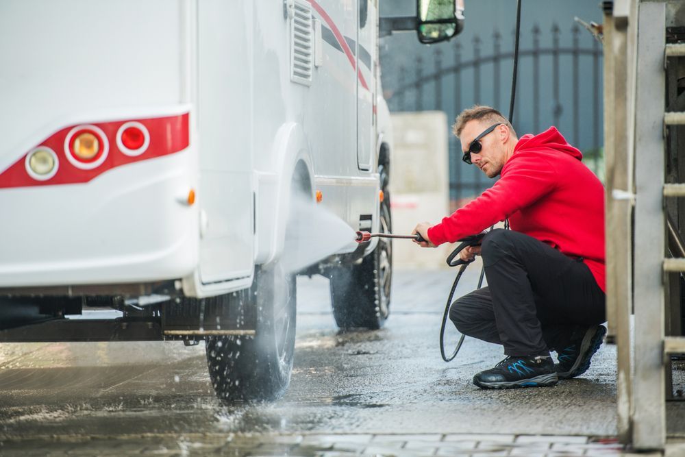 Man in a red jacket washing a white van with a hose at a car wash