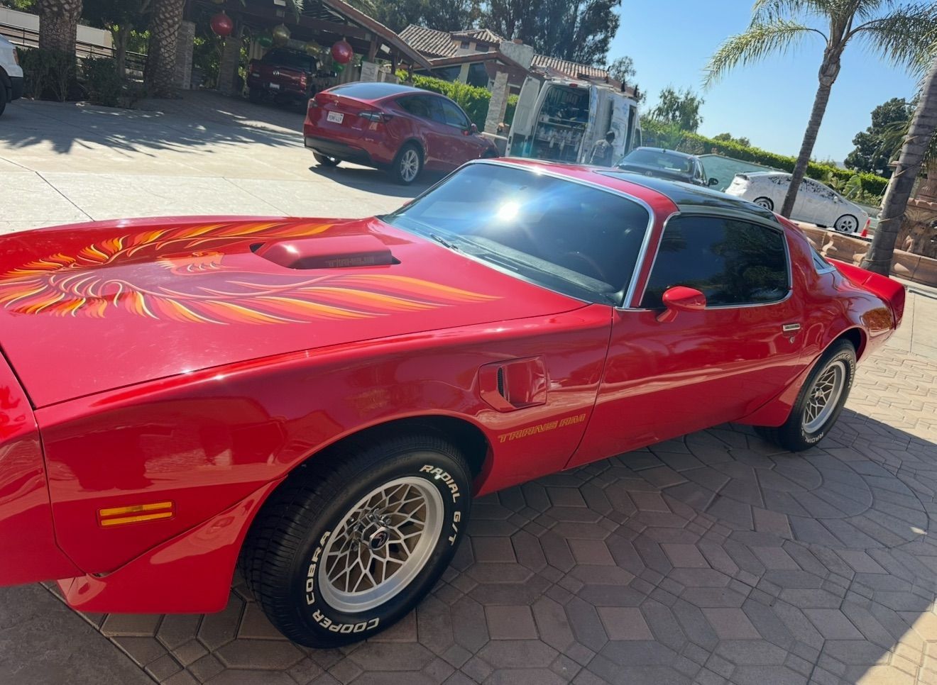 Red classic sports car parked on a sunlit driveway near a house, with another red car in the background