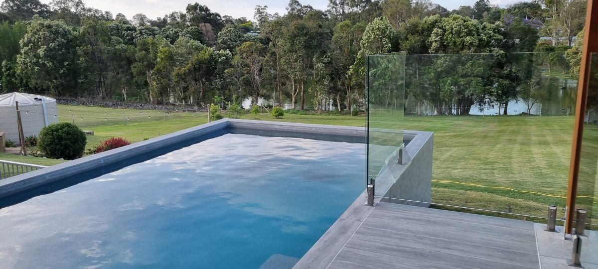 A Large Swimming Pool Is Surrounded By A Glass Fence And Trees — CJB Pools in Pie Creek, QLD