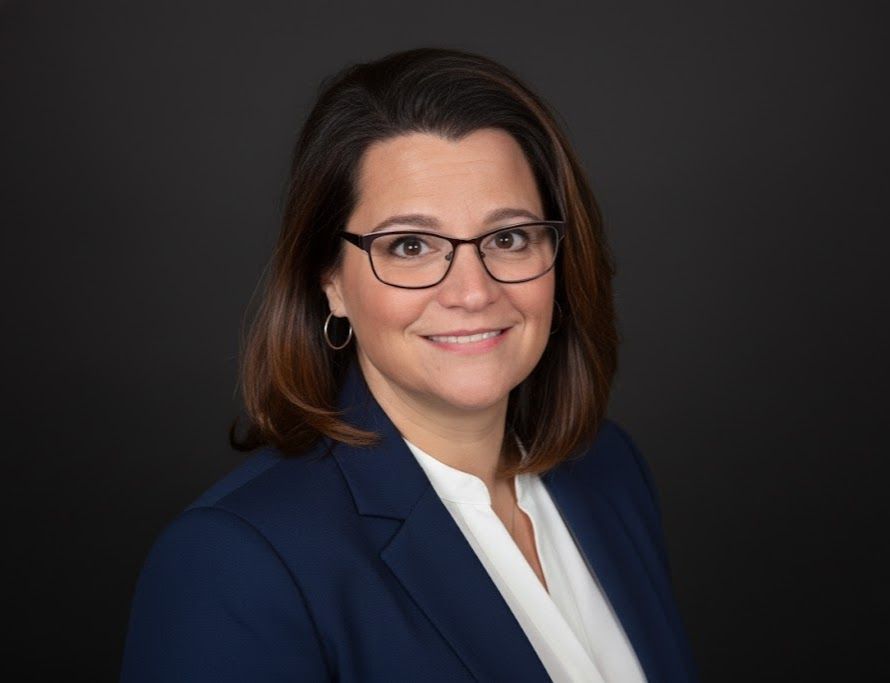 Portrait of a smiling person in glasses, wearing a navy blazer and white blouse on a dark background