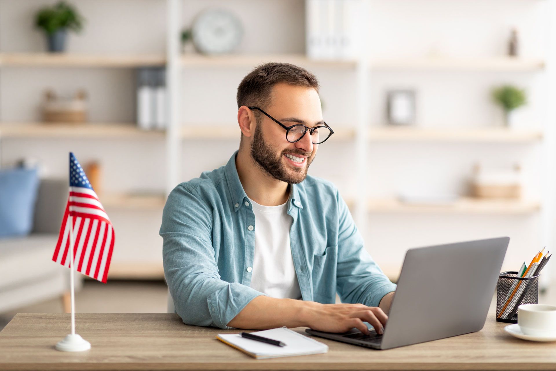 Man typing on a laptop at a desk with a small American flag in a bright home office