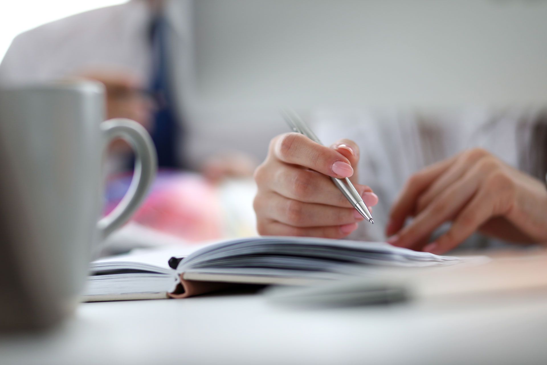 Hands writing in an open notebook at a desk with a coffee mug in the foreground