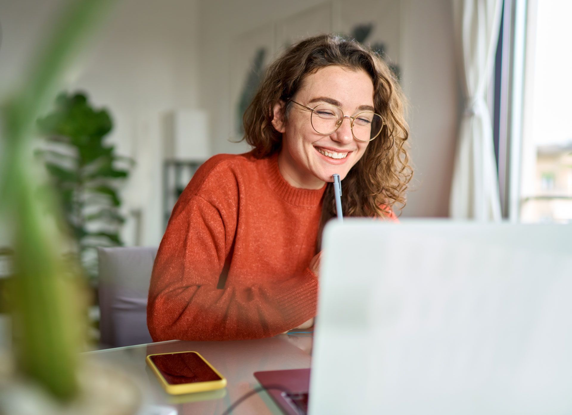 Smiling woman in a red sweater using a laptop at a desk with a phone nearby