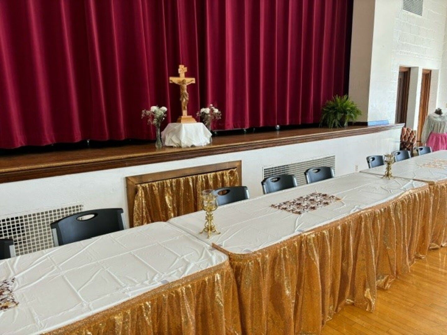 A decorated stage with a red curtain backdrop features a wooden crucifix and floral arrangements.