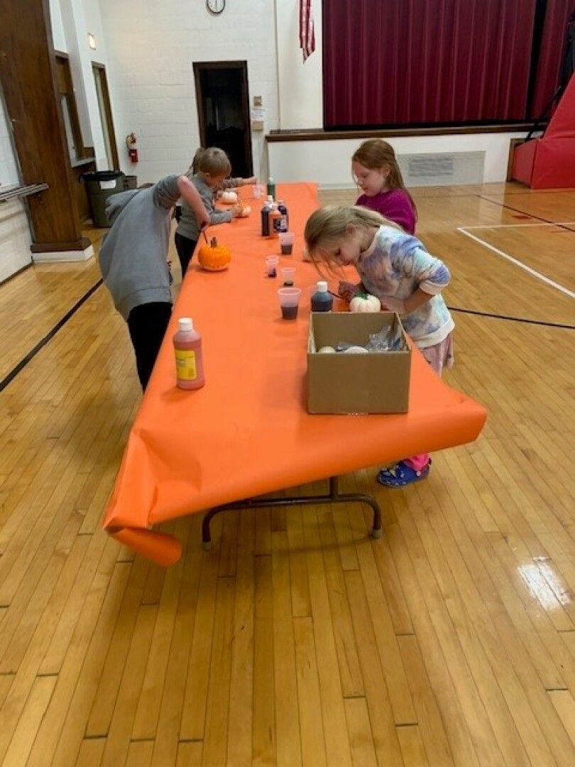 Three children paint pumpkins on an orange tablecloth-covered table in a gymnasium.