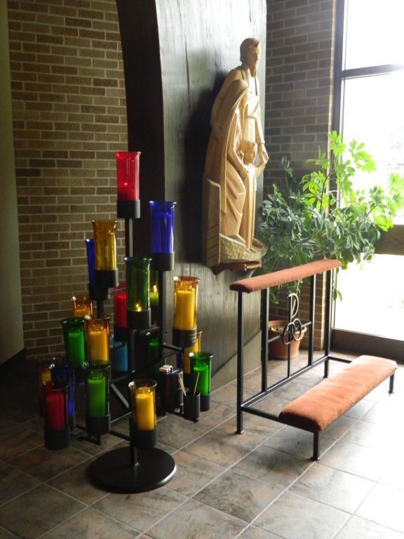 A serene chapel corner featuring a wooden religious statue, vibrant multicolored votive candles.