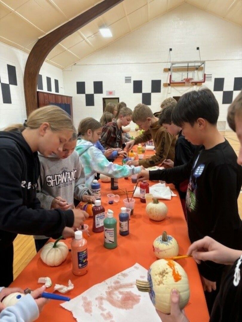 Children gather around a table in a gymnasium, painting small pumpkins.