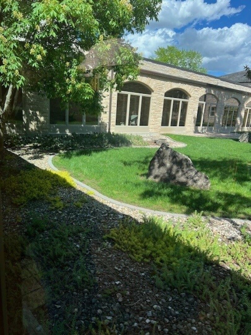 A serene courtyard with a stone building featuring arched windows, lush green grass, a large boulder, and a tree casting shadows.