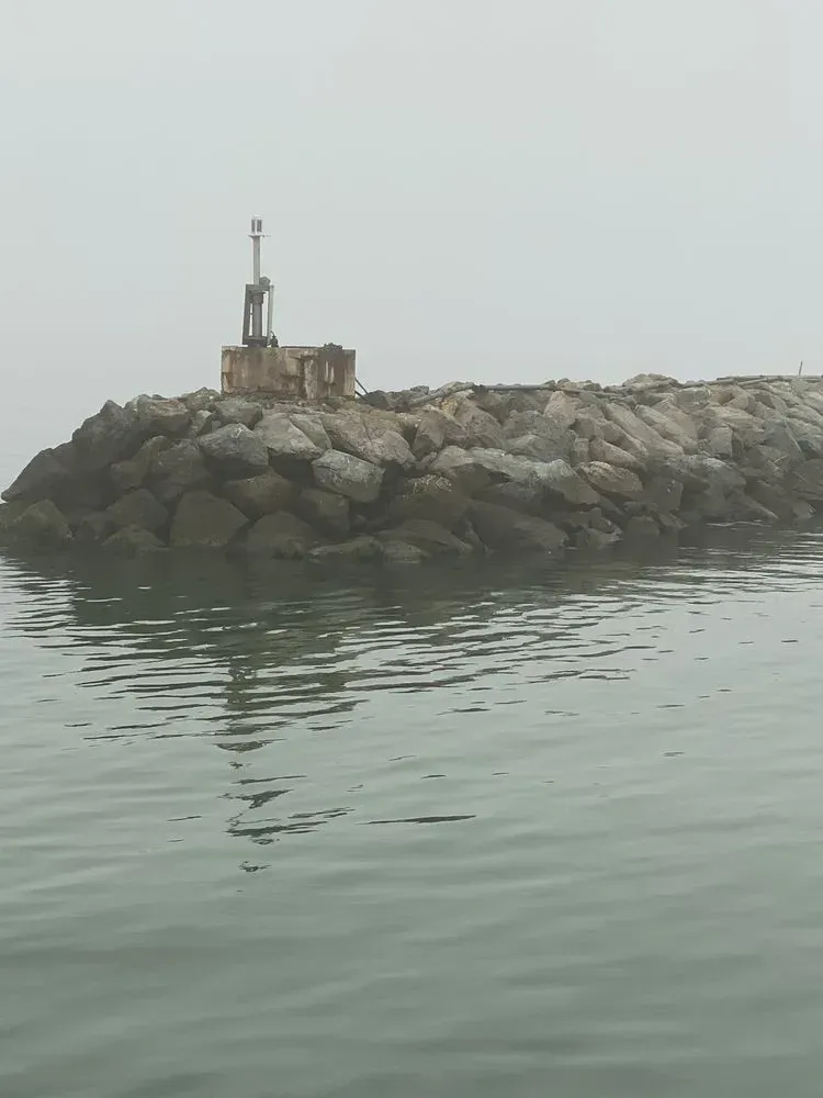 Rock jetty with a small structure and a beacon under a foggy gray sky. Calm, green-tinted water.
