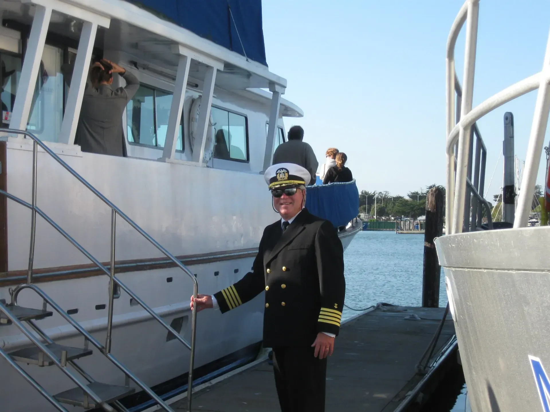 Man in captain's uniform stands on dock next to a white boat, holding a cane. People on boat in background.