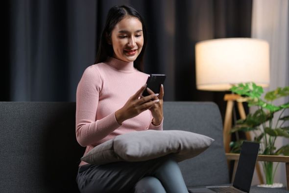 Woman in a pink sweater smiling at a smartphone on a couch with a pillow, beside a lit lamp and laptop.