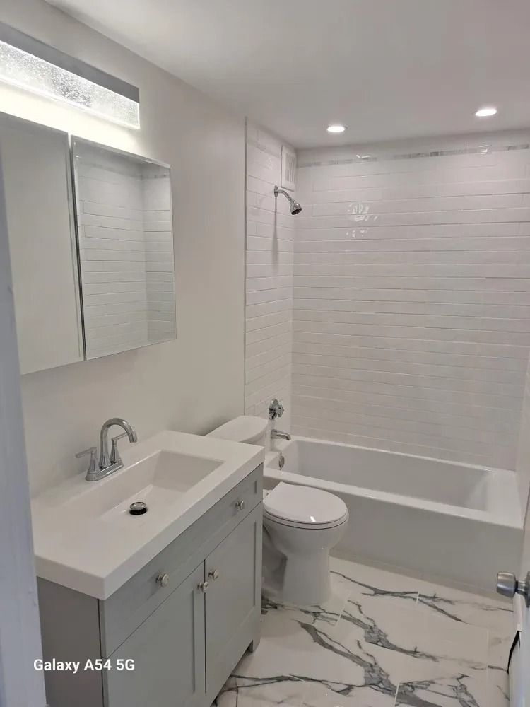 Modern white bathroom with sink, toilet, bathtub, and marble-pattern tile floor.