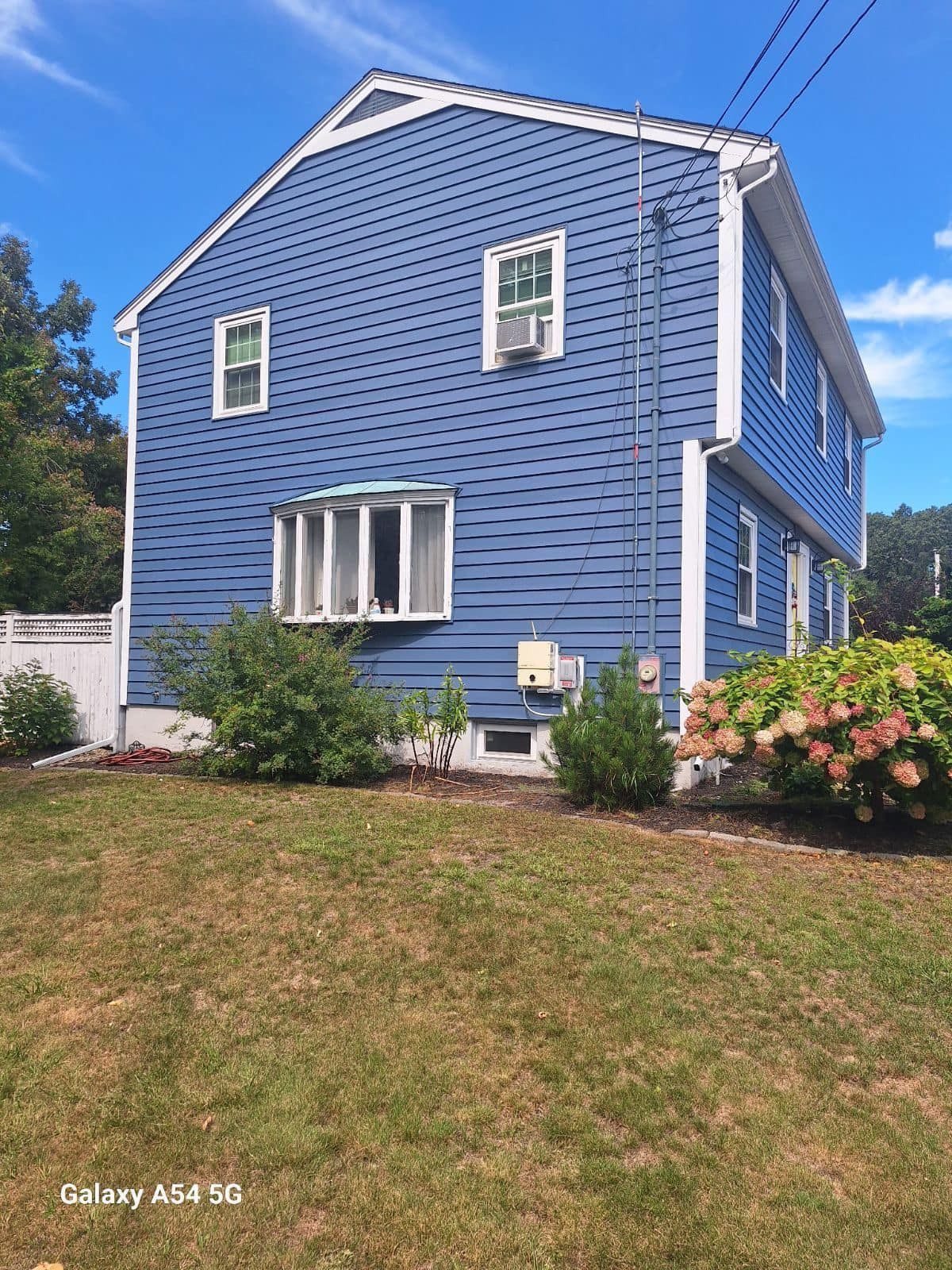 Blue two-story house with white trim, bay window, and shrubs on a sunny grassy lawn
