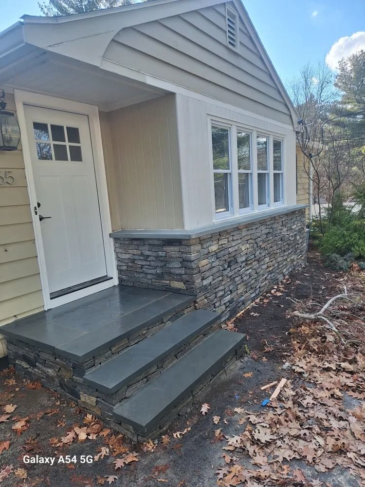 Small beige house exterior with stone base, white door, front steps, and large windows.