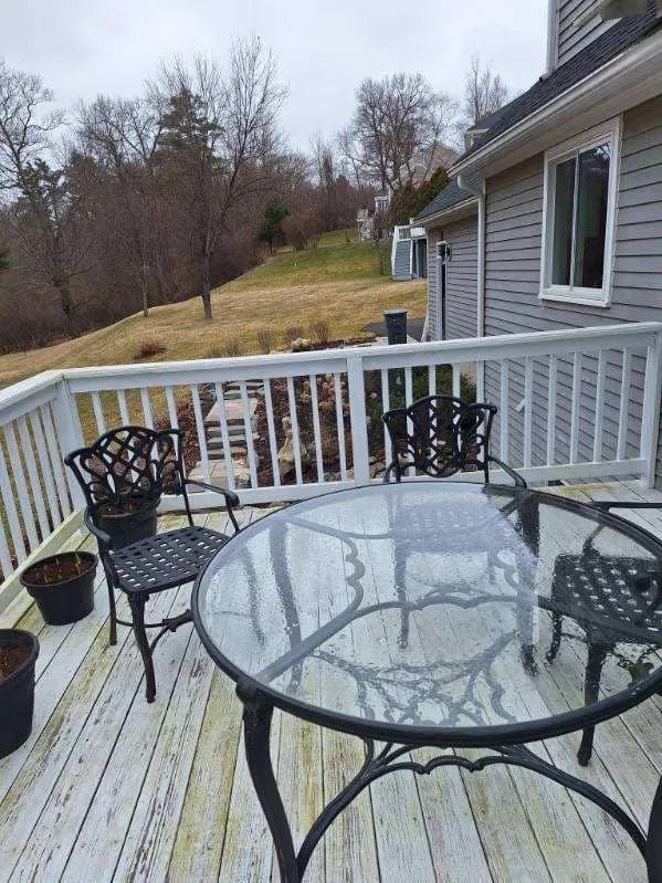 Glass patio table with black chairs on a deck beside a house, overlooking a grassy hillside and trees.