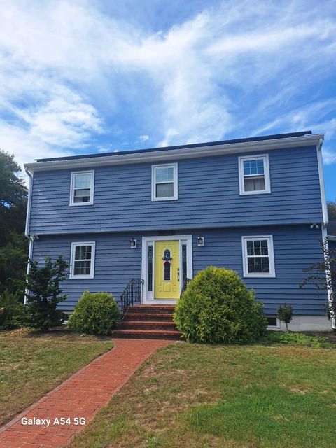 Blue two-story house with white trim, yellow front door, red walkway, and green shrubs under a cloudy sky