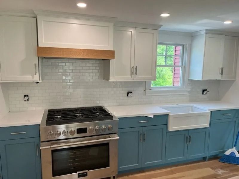 Modern kitchen with blue lower cabinets, white upper cabinets, stainless stove, and a farmhouse sink by a window.