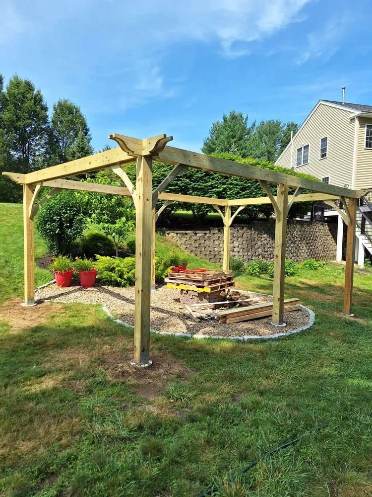 Wooden pergola over a circular fire pit in a grassy backyard beside a house.