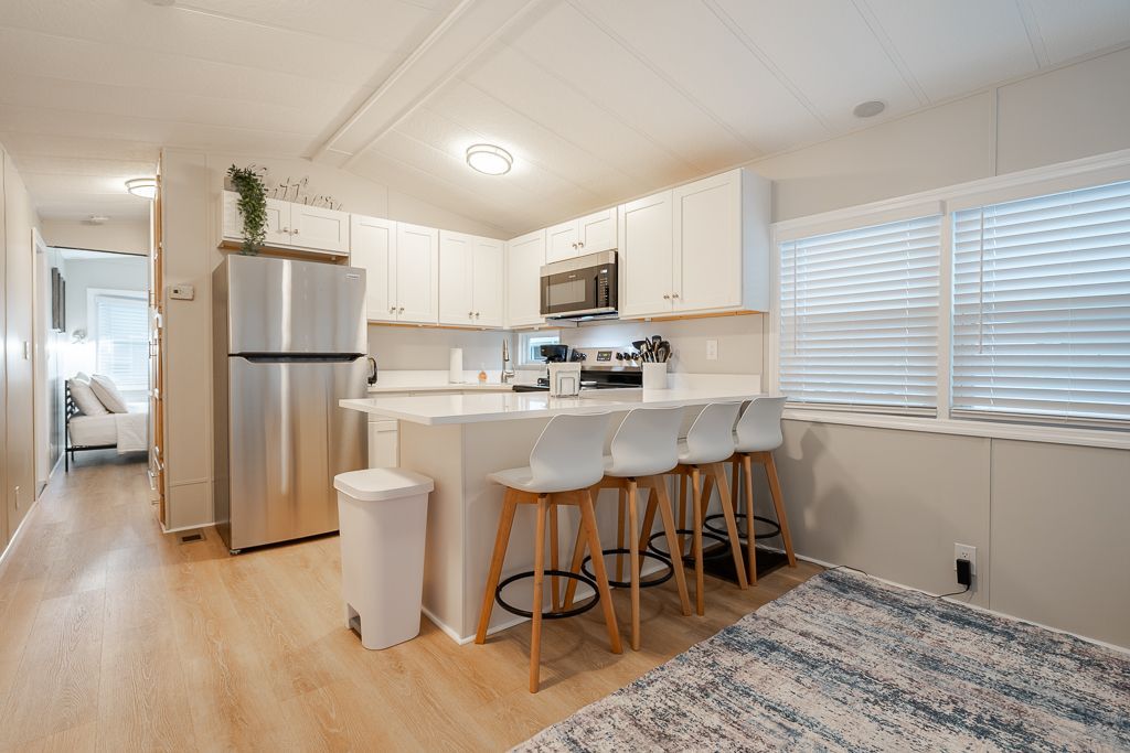 A bright kitchen featuring white cabinets, stainless steel appliances, and a breakfast bar with four white bar stools.