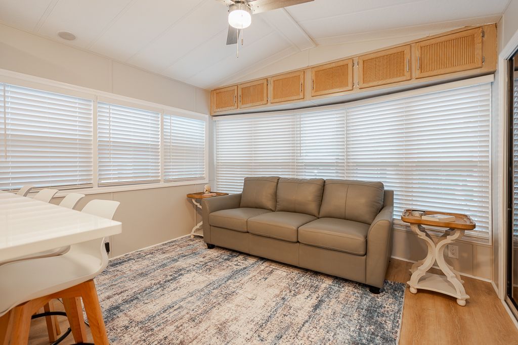 A bright living area with a tan sofa, patterned area rug, a side table, and windows covered by horizontal blinds.