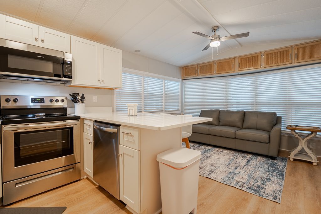A kitchen with white cabinets, stainless steel appliances, and a breakfast bar next to a living room with a grey sofa.