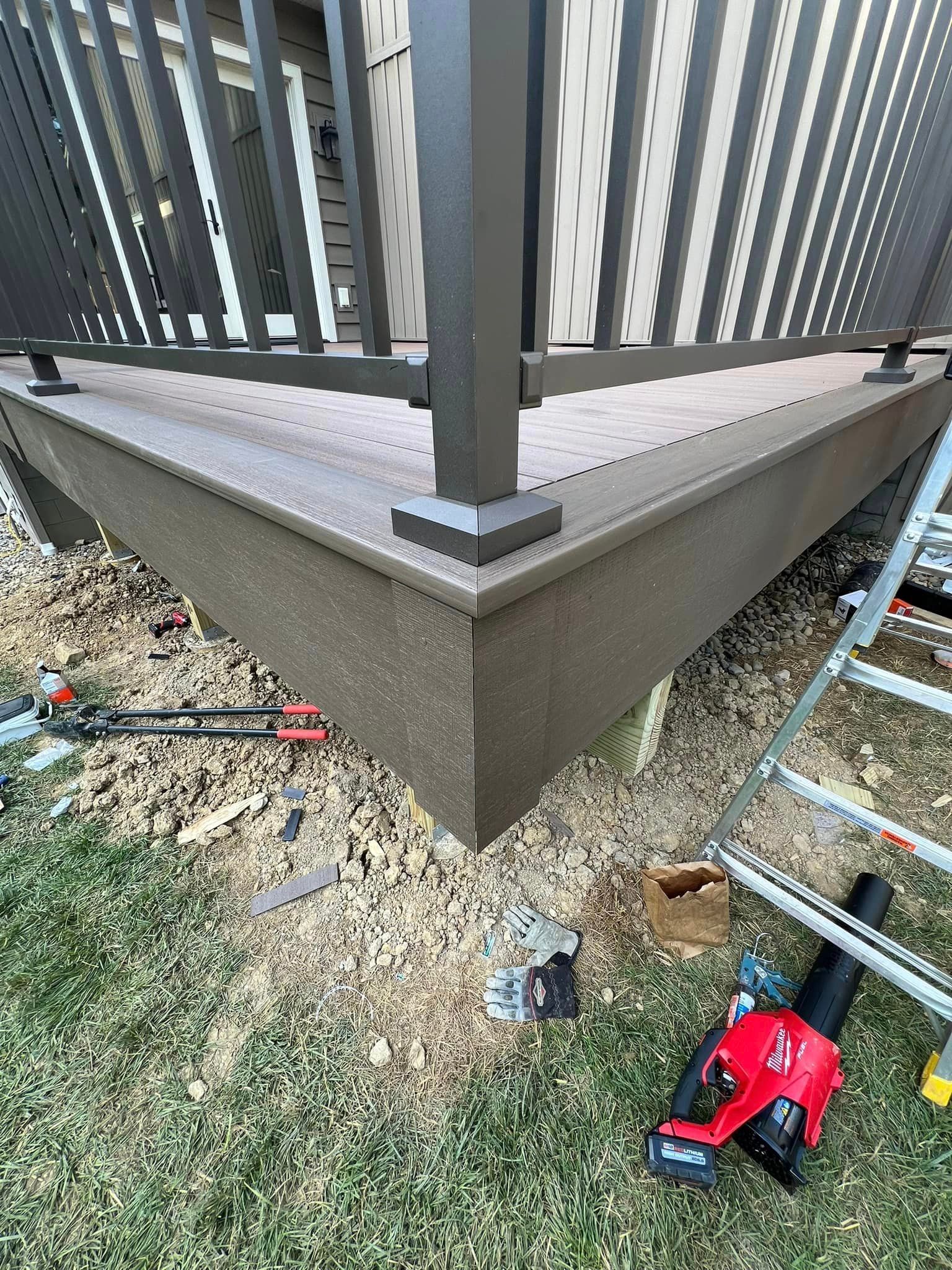 A close-up of a brown composite deck corner with a black railing, set above a yard with tools and a ladder nearby.