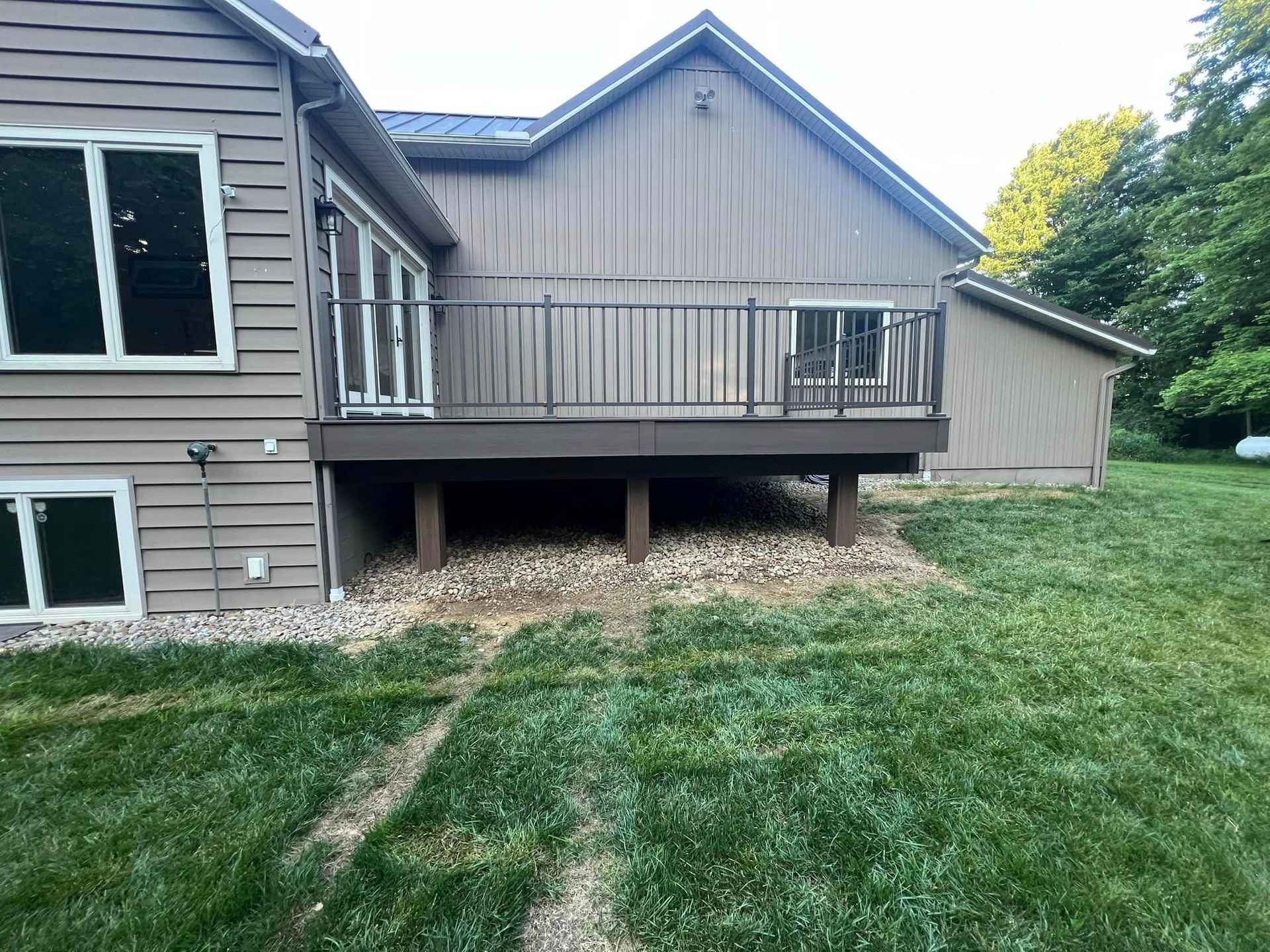 A brown house with a raised deck, metal railings, and a gravel base, set against a green lawn and trees.
