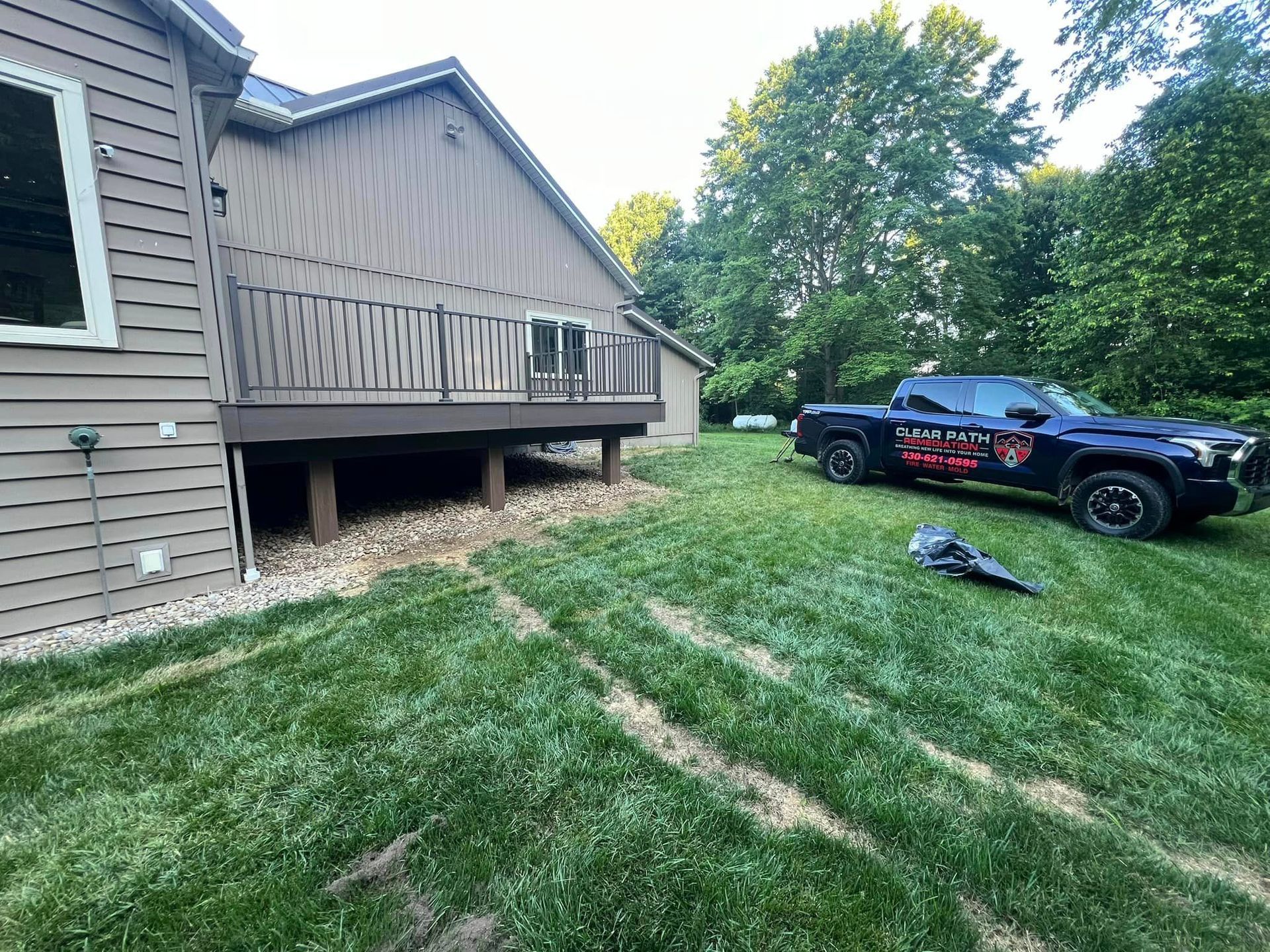 A tan house with a wooden deck overlooks a grassy lawn, with a dark pickup truck parked nearby.