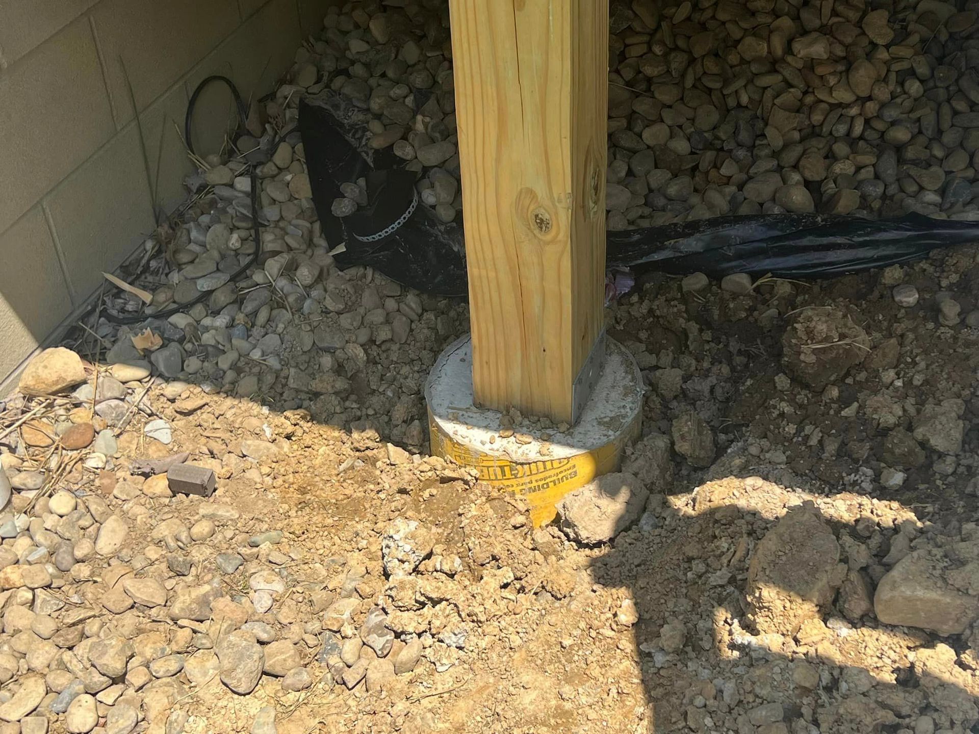 A wooden deck post supported by a cylindrical concrete footer installed in a gravel and soil yard.