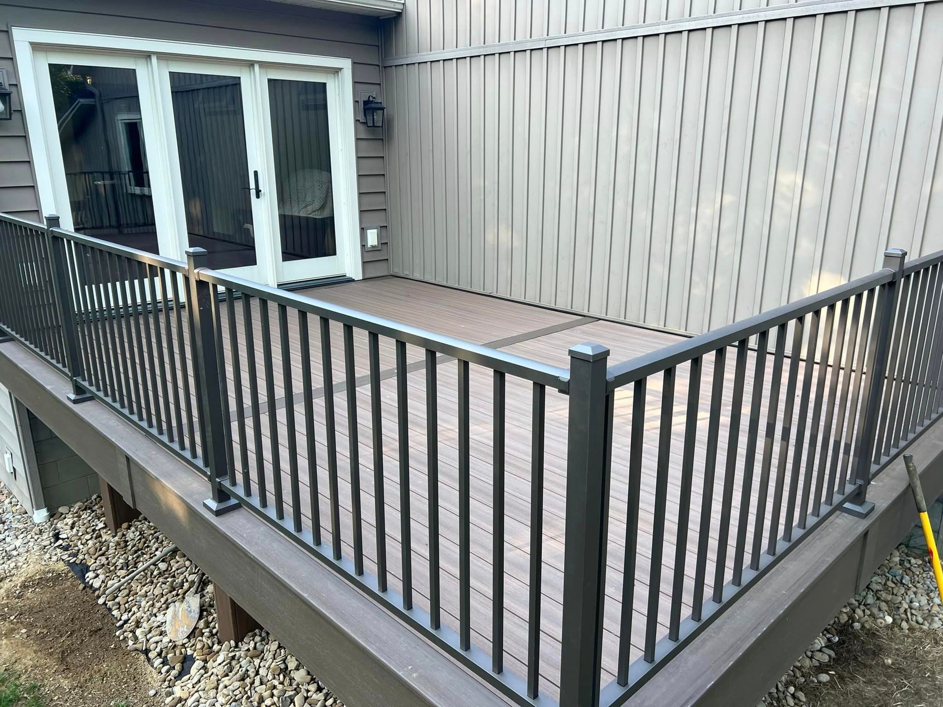 A low-angle view of a brown composite deck with black metal railings, leading to glass double doors on a tan house.
