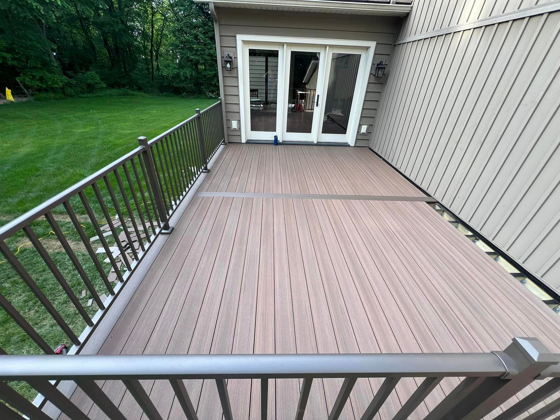 A light brown composite deck with a metal railing, overlooking a green lawn next to a house with glass doors.
