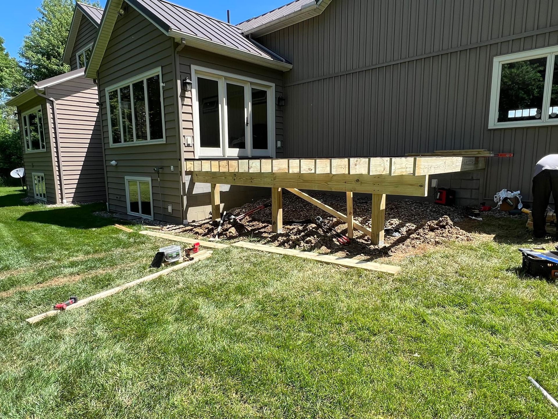 A wooden deck frame under construction attached to the back of a tan siding house with a grassy yard.