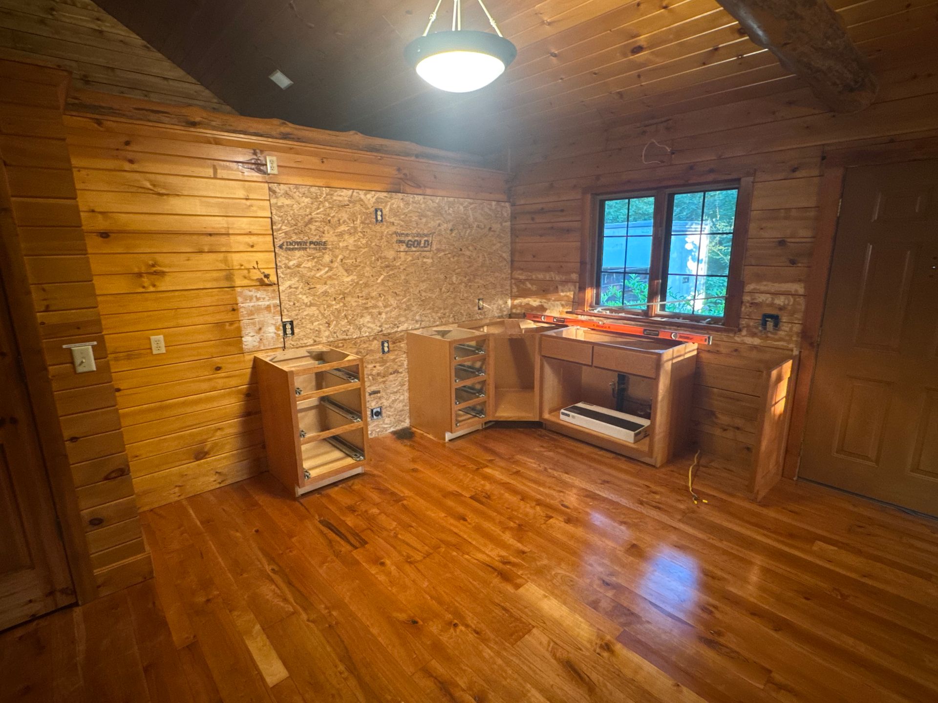 An unfinished kitchen with wooden cabinets and OSB wall panels inside a room with rustic wood walls and hardwood floors.