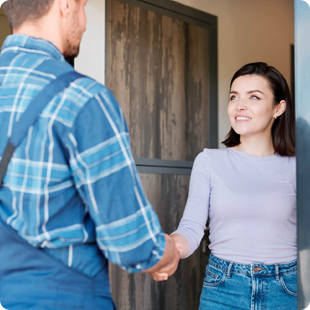 A worker in a blue plaid shirt and overalls shakes hands with a person in a light purple long-sleeved shirt by a doorway.
