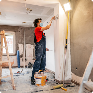 A worker in a red shirt and blue overalls uses a trowel to smooth plaster onto an indoor wall during a renovation.