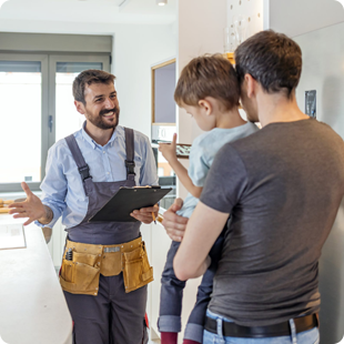 A professional in work coveralls and a tool belt speaks with a person holding a child in a home kitchen setting.