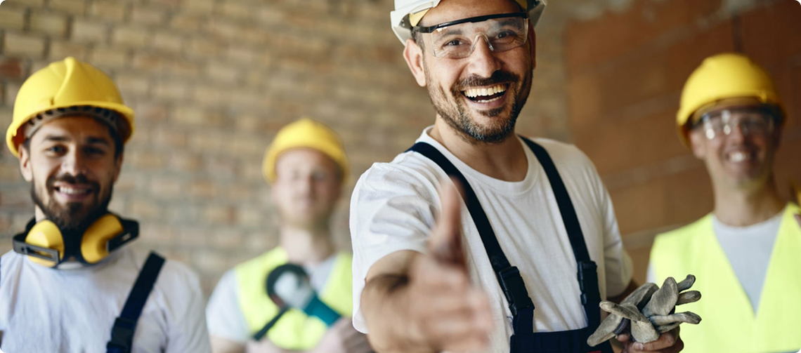 Four construction workers in hard hats and safety gear smiling in a brick building; one extends a hand for a handshake.