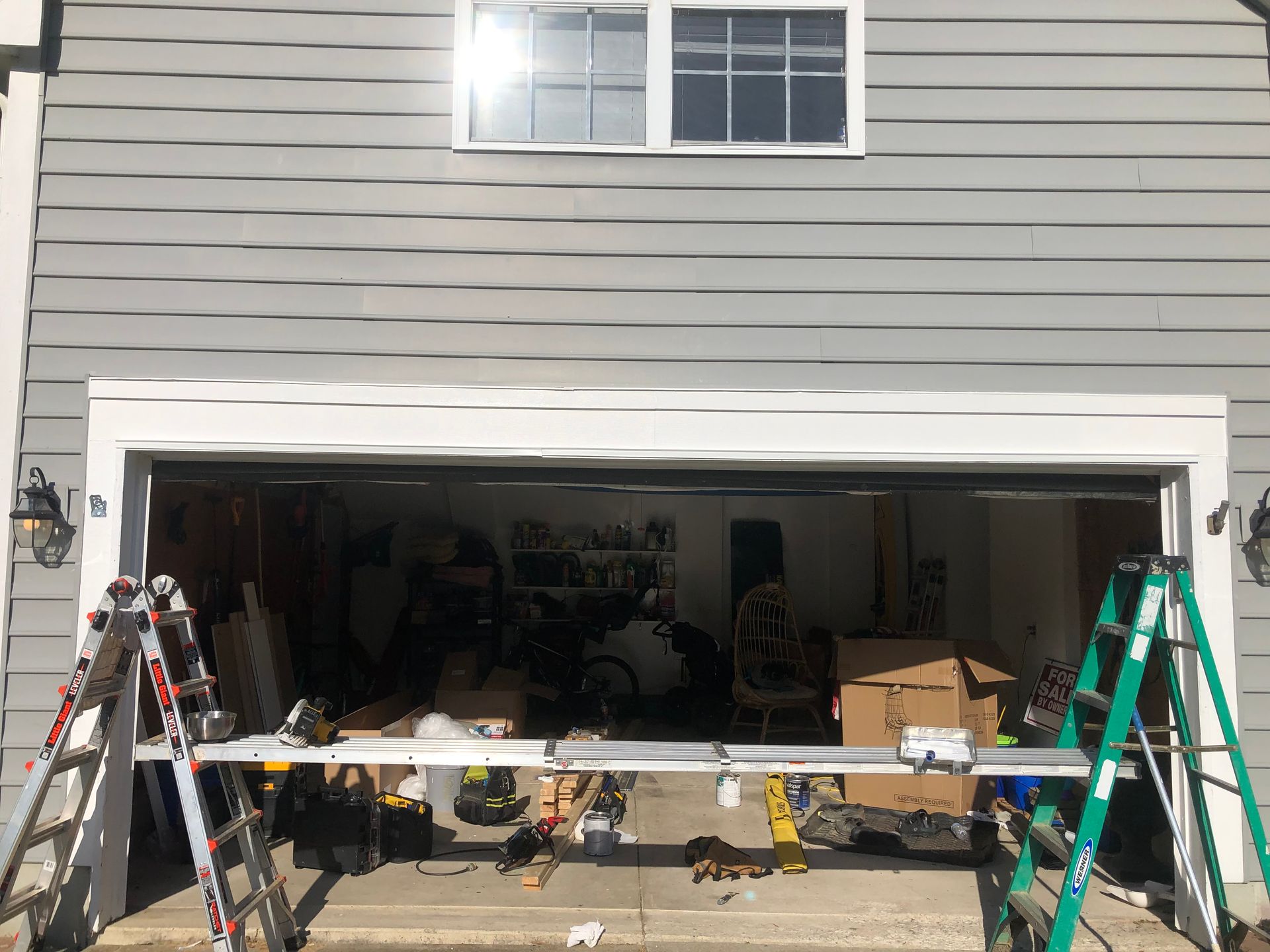 Two ladders stand in front of an open residential garage, supporting a long metal plank across the doorway.