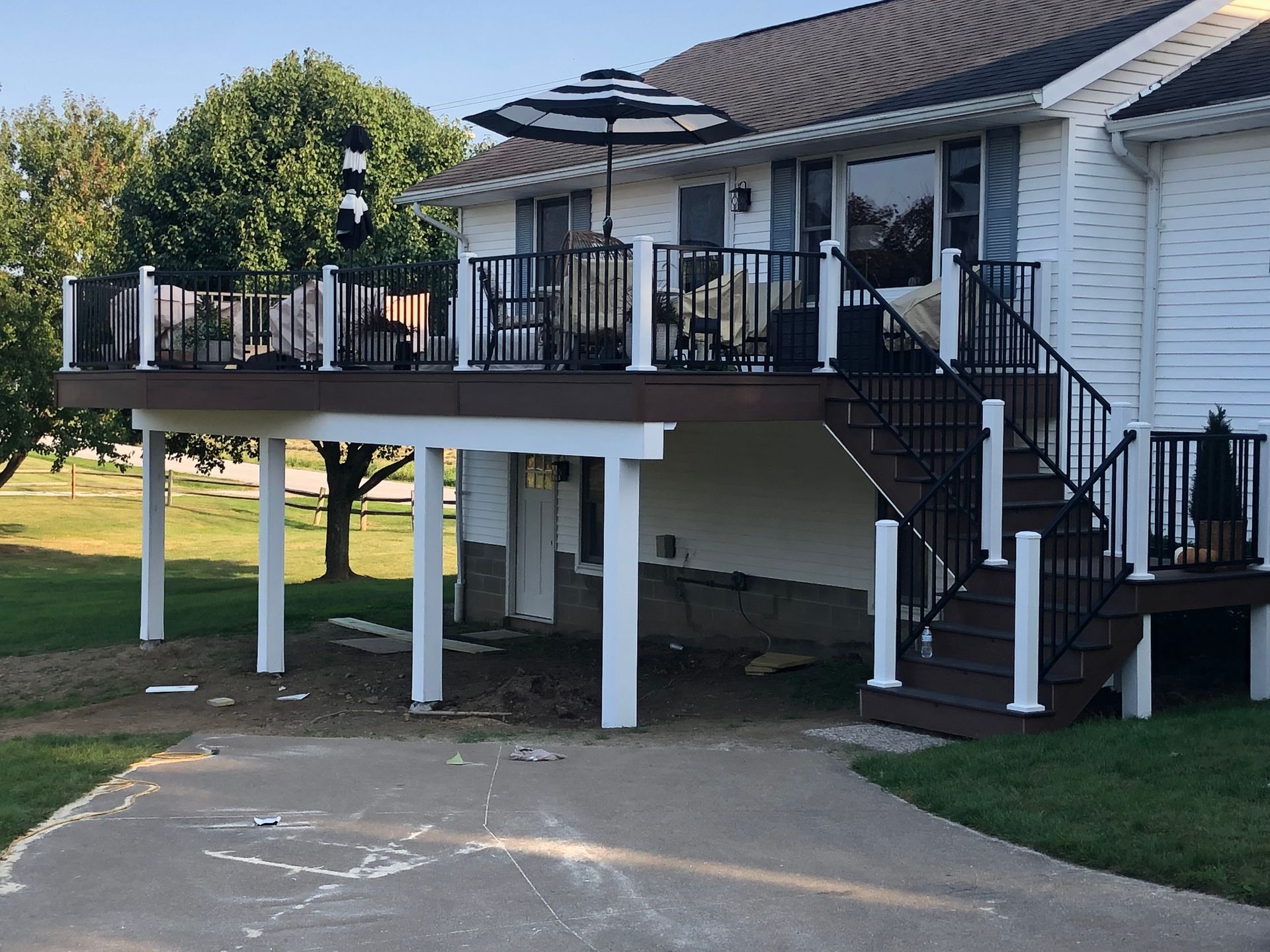 A brown wooden deck with black railings and stairs, supported by white posts, attached to the back of a white house.