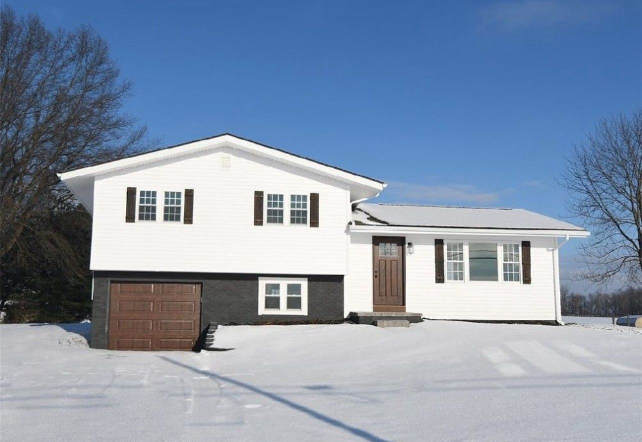 A two-story white house with a dark brown garage door, wooden front door, and black shutters, surrounded by snow.