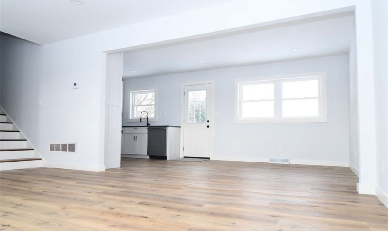 A bright, open-concept living space with light-colored flooring, white walls, stairs, and a view into a small kitchen.
